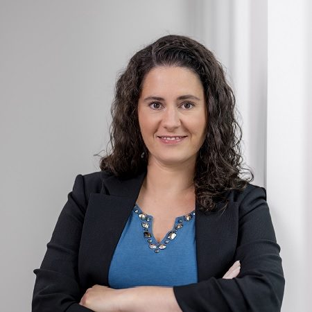 Die abgebildete Person trägt eine hellblaue Bluse mit Zierdetails am Ausschnitt und einen schwarzen Blazer. Die Person steht mit verschränkten Armen vor einem weißen Hintergrund.