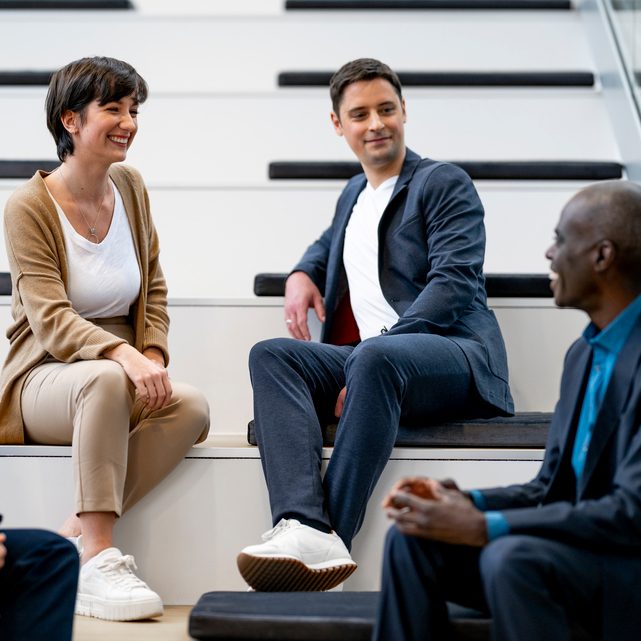 Das Bild zeigt eine Gruppe von Personen, die in einem professionellen oder geschäftlichen Umfeld auf Treppen sitzen und sich miteinander unterhalten. Eine Frau und zwei Männer sind sichtbar, wobei sie lässige oder geschäftliche Kleidung tragen. Die Szene findet vermutlich in einem modernen Büro oder Konferenzraum statt.