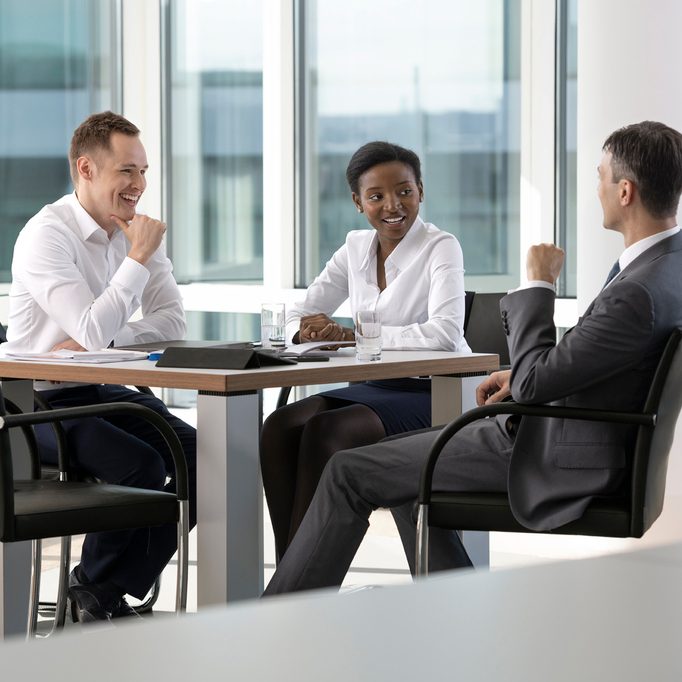 Das Bild zeigt eine Gruppe von drei Personen in formeller Kleidung, die an einem Tisch in einem modernen Büroumfeld eine Besprechung abhalten. Das Büro verfügt über große Fenster, die viel Tageslicht hereinlassen, und die Personen scheinen in einem Gespräch vertieft zu sein.