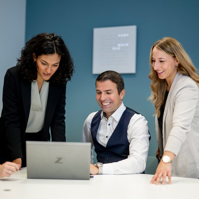 Das Bild zeigt eine Gruppe von Personen, die in einem professionellen Büro mit einem Laptop interagieren. Sie scheinen eine Diskussion oder Zusammenarbeit zu führen.