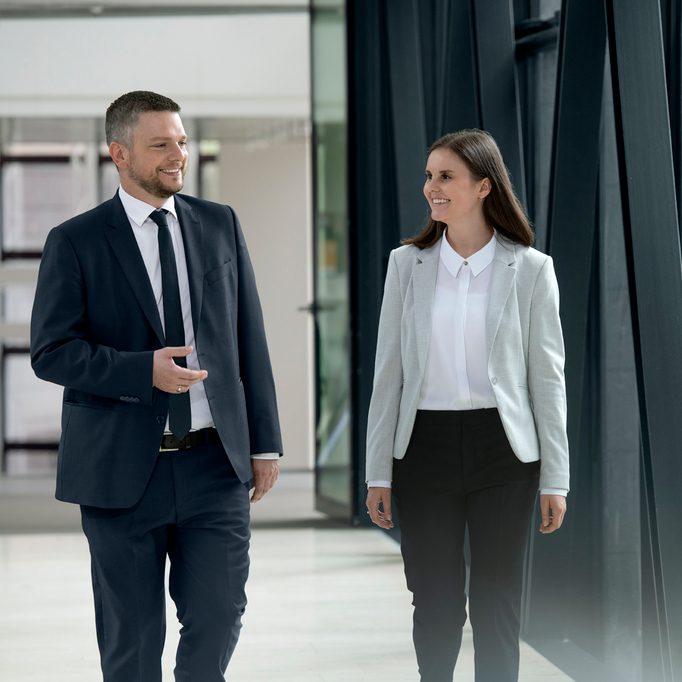 Das Bild zeigt zwei Personen in formeller Kleidung in einem modernen Bürogebäude oder einer Geschäftsumgebung. Sie scheinen einem Gespräch nachzugehen, möglicherweise im Rahmen eines Geschäftstreffens oder einer Besprechung.