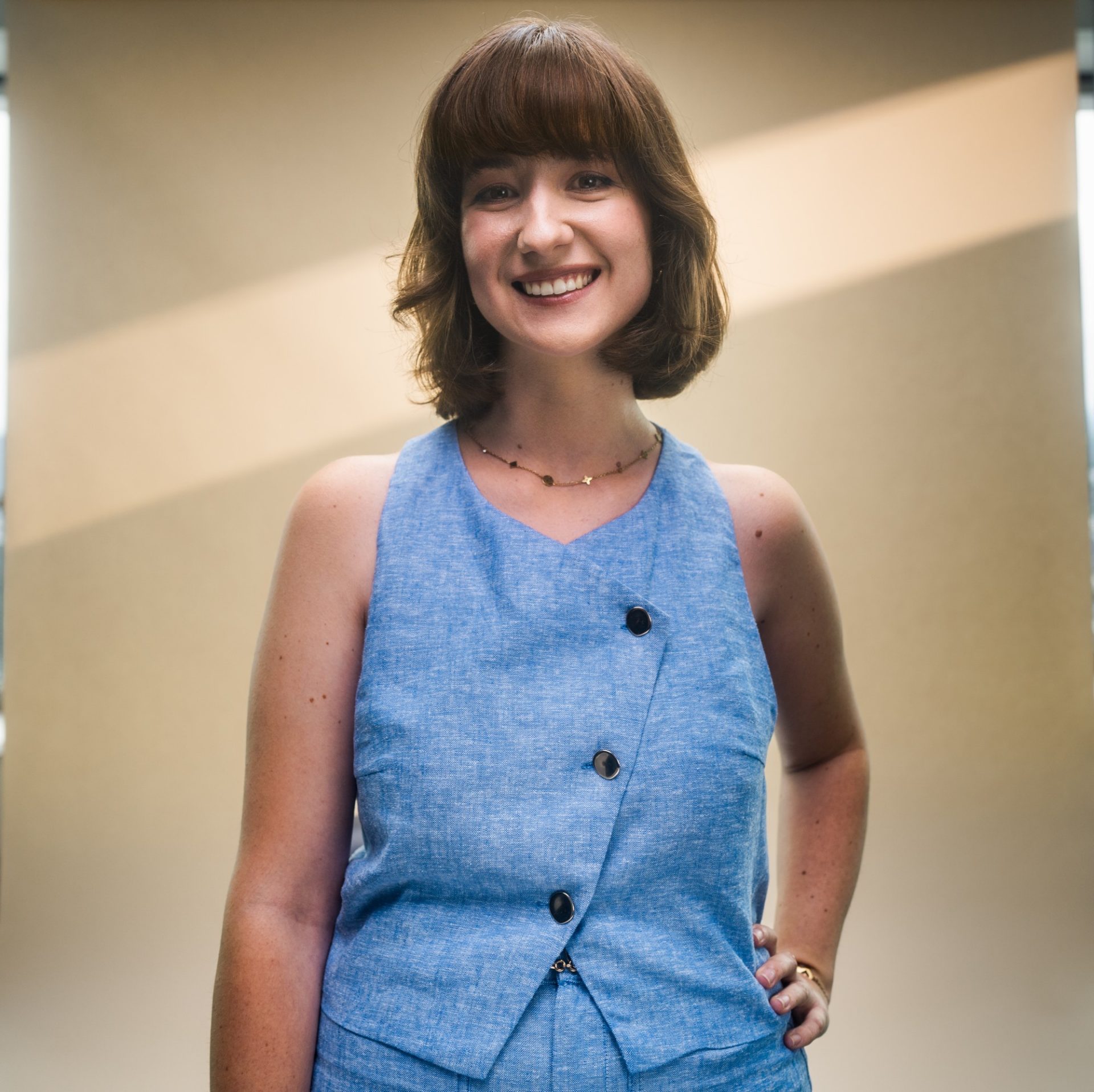 Eine Person, die ein ärmelloses Oberteil mit Knöpfen trägt. Das Hintergrundbild zeigt eine helle, neutrale Wand mit einem diagonalen Lichtstrahl.