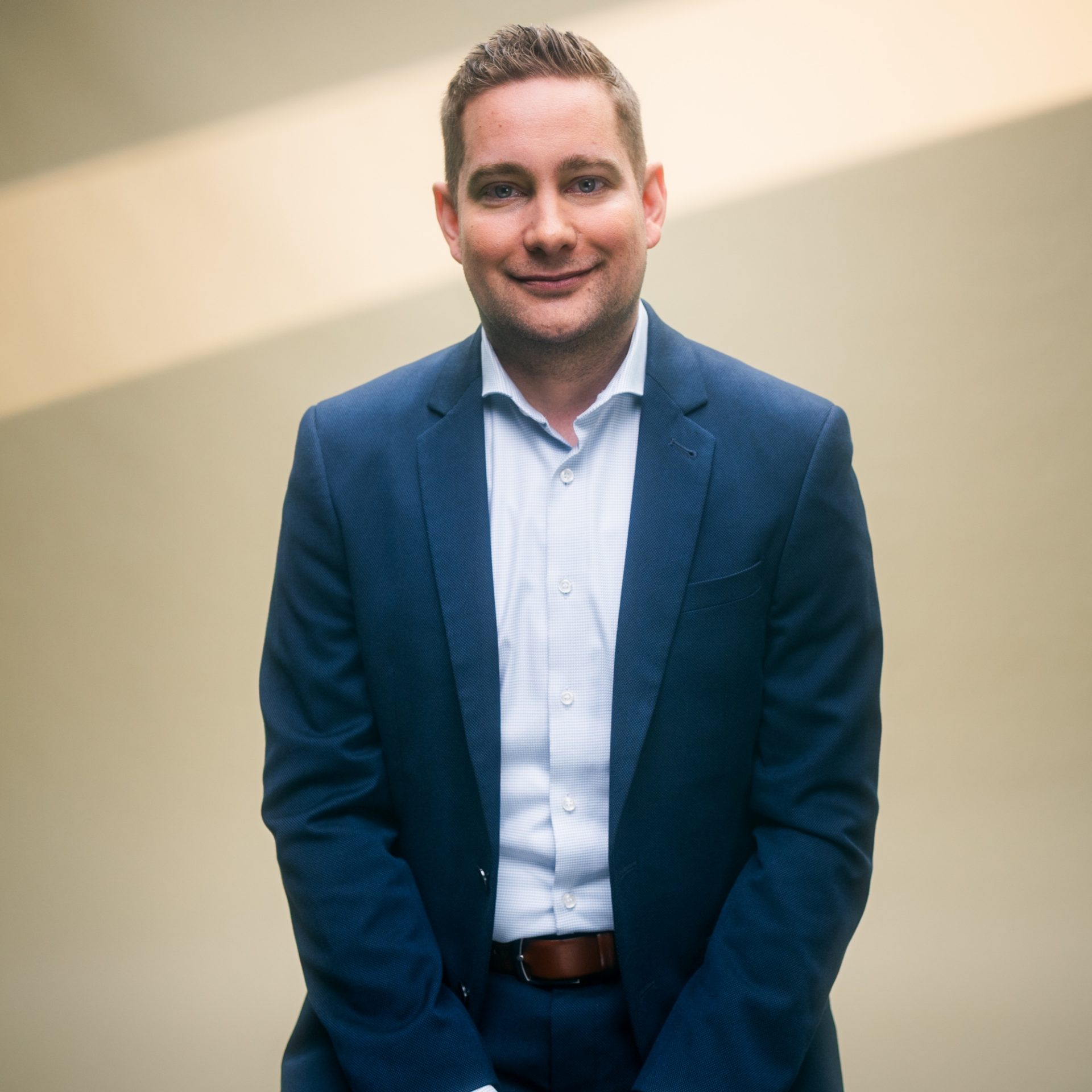 Eine Person in einem dunklen Anzug und weißem Hemd steht vor einem neutralen Hintergrund.