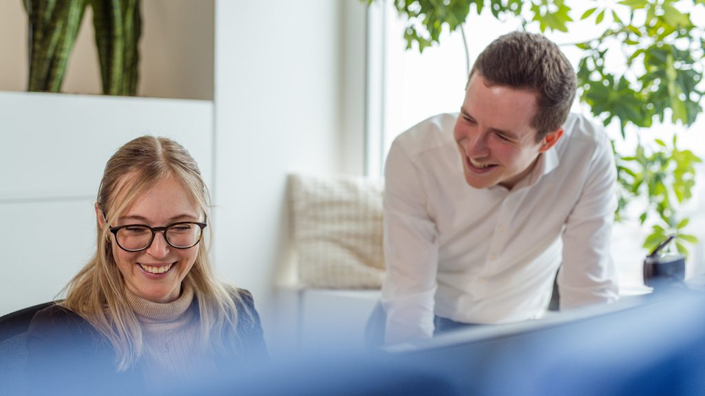 Ein Büro mit zwei Personen vor einem Bildschirm, wobei eine Person steht und die andere sitzt. Es gibt Pflanzen im Hintergrund und es wirkt wie eine entspannte und professionelle Büroumgebung.