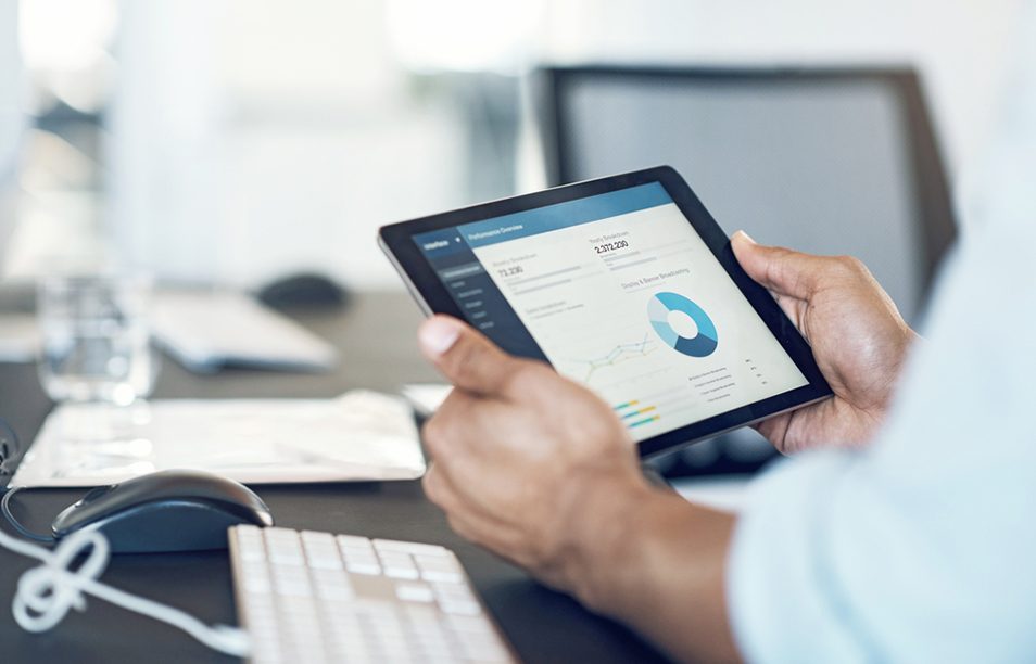 Das Bild zeigt eine Person, die ein Tablet hält und darauf eine graphische Darstellung von Daten oder Statistiken betrachtet. Im Vordergrund sind Teile einer Tastatur und einer Maus sichtbar, was darauf hindeutet, dass die Person an einem Arbeitsplatz oder in einem Büro sitzt. Das Bild vermittelt den Eindruck von technologischem Fortschritt und effizienter Datenanalyse, die in modernen Arbeitsumgebungen eingesetzt wird.