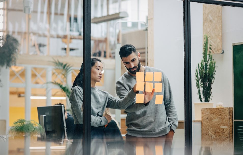 Das Bild zeigt eine Umgebung in einem Büro oder Arbeitsbereich, in der zwei Personen zusammenarbeiten. Sie wirken konzentriert und scheinen etwas auf die Post-It-Notizen an der Glasscheibe zu schreiben oder zu besprechen. Im Hintergrund sieht man einen Raum mit verschiedenen Möbelstücken und Pflanzen, die eine freundliche und inspirierende Atmosphäre schaffen.