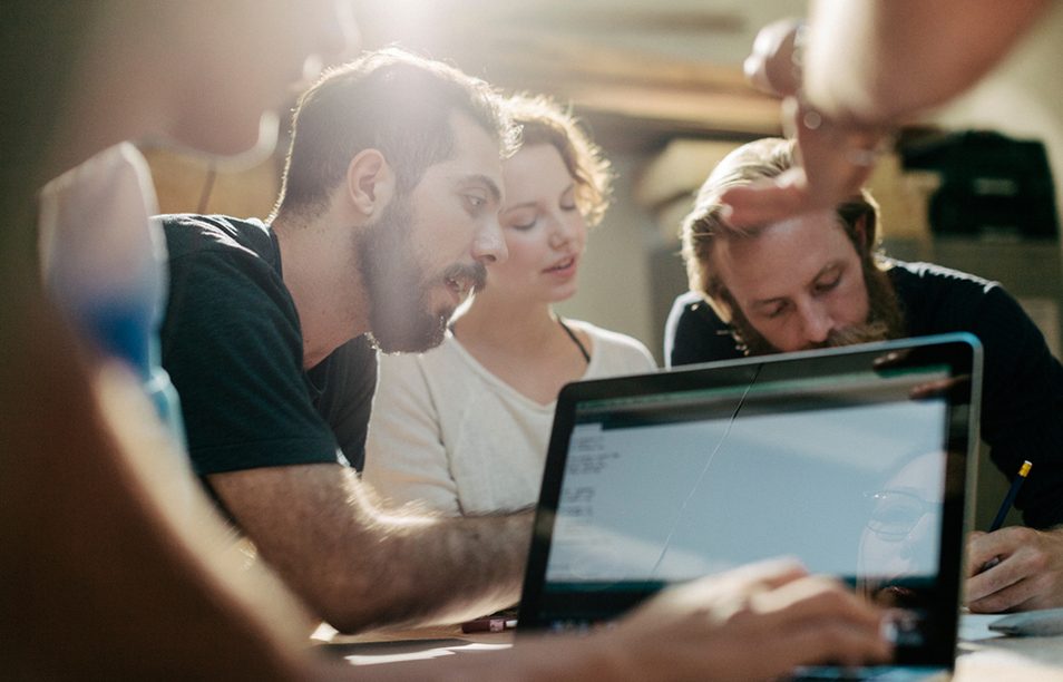 Das Bild zeigt eine Gruppe von Personen, die um einen Tisch herum sitzen und gemeinsam an einem Laptop arbeiten. Einige von ihnen scheinen etwas zu erklären oder zu diskutieren, während sie auf den Bildschirm schauen. Es handelt sich um eine Szene im Rahmen eines Team- oder Gruppenarbeitstreffens in einem Büro oder Arbeitsraum.