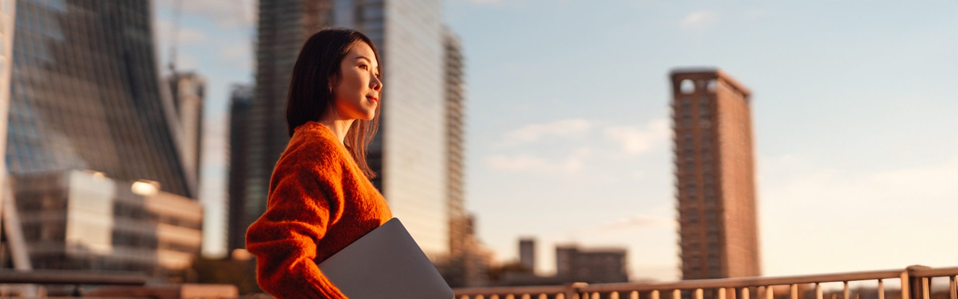 Das Bild zeigt eine Frau, die auf einem Balkon oder einer Terrasse in einer städtischen Umgebung steht. Die Sonne scheint und taucht die Szene mit einem warmen Licht. Die Frau trägt einen orangenen Pullover und hält einen Laptop. Im Hintergrund sind moderne Wolkenkratzer zu sehen, was auf eine Großstadt hinweist. Das Bild vermittelt einen Moment des Nachdenkens oder der Entspannung inmitten städtischen Treibens.