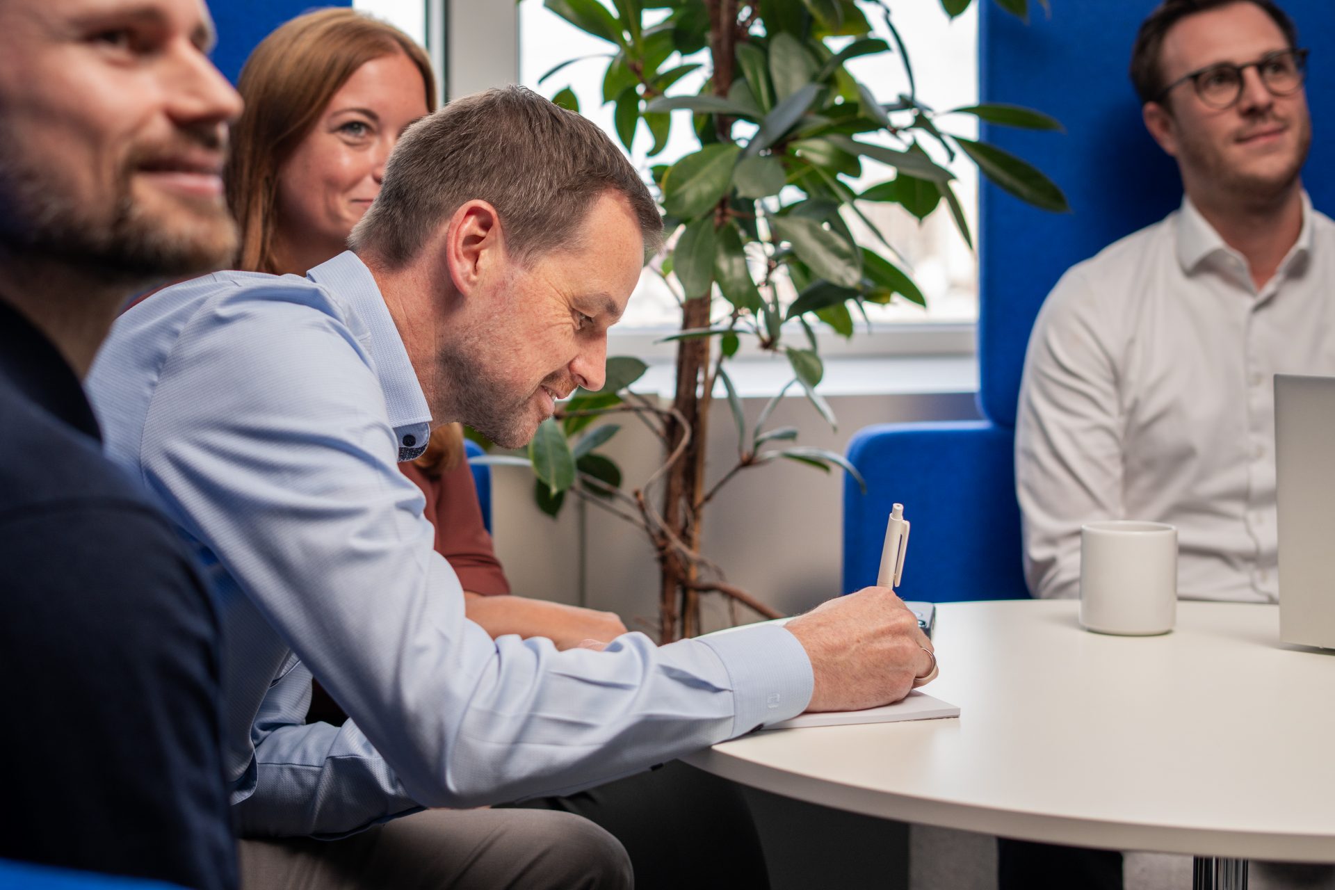 Ein Gruppenmeeting oder eine Teambesprechung in einem Büro, bei dem mehrere Personen um einen Tisch versammelt sind. Eine Person notiert etwas auf einem Block. Im Hintergrund ist eine Pflanze und ein Fenster sichtbar, und auf dem Tisch steht eine weiße Tasse. Das Setting deutet auf eine formelle Umgebung hin, möglicherweise in einem Konferenzraum.