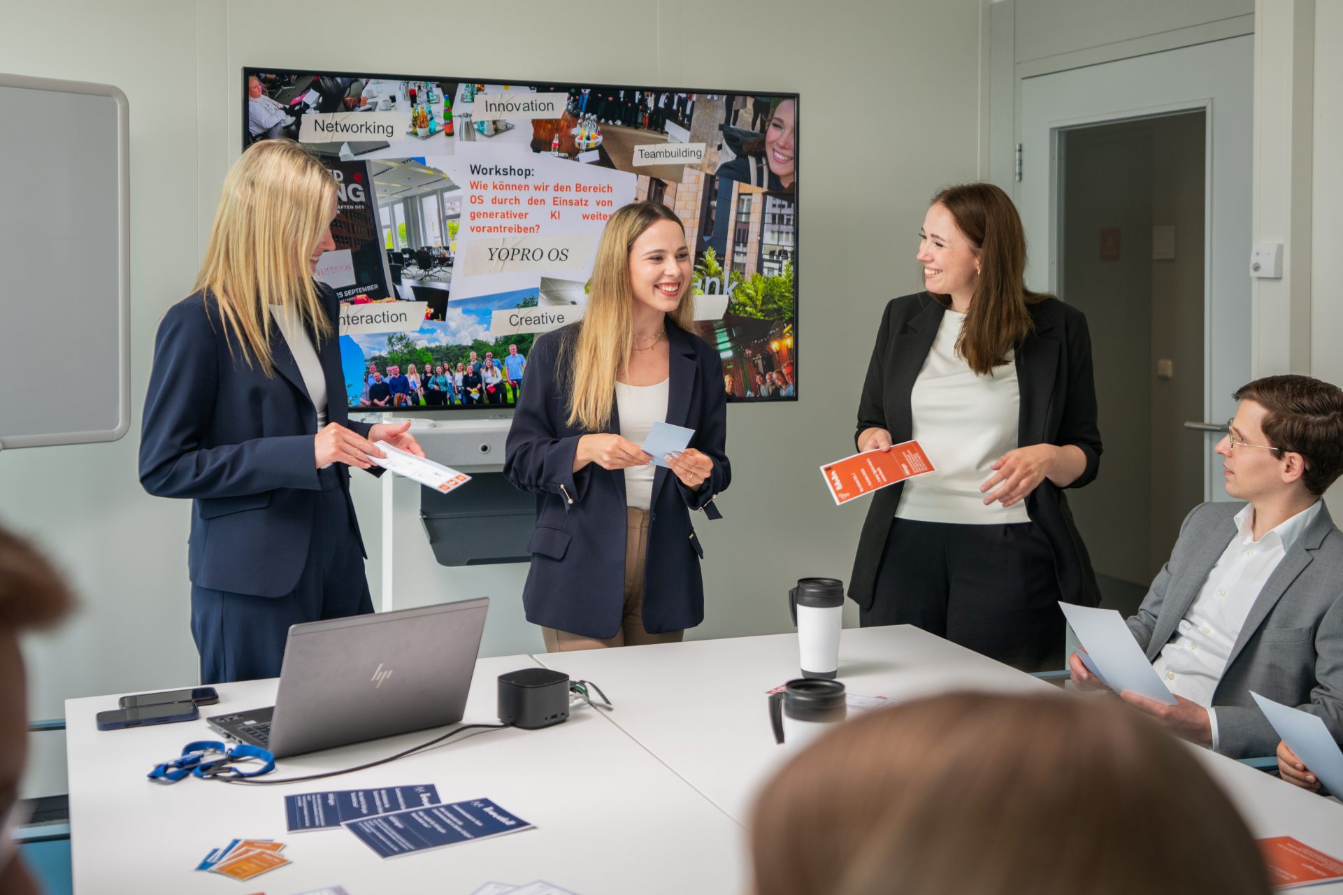 Das Bild zeigt ein formelles Geschäftstreffen oder eine Präsentation in einem Büroraum. Drei Personen sind in professioneller Kleidung und stehen vor einem Tisch mit einem Laptop, Dokumenten und Kaffeetassen, während sie anscheinend diskutieren oder präsentieren. Im Hintergrund ist ein Bildschirm mit verschiedenen Bildern und Texten, der möglicherweise Themen oder Informationen zum Meeting zeigt.
