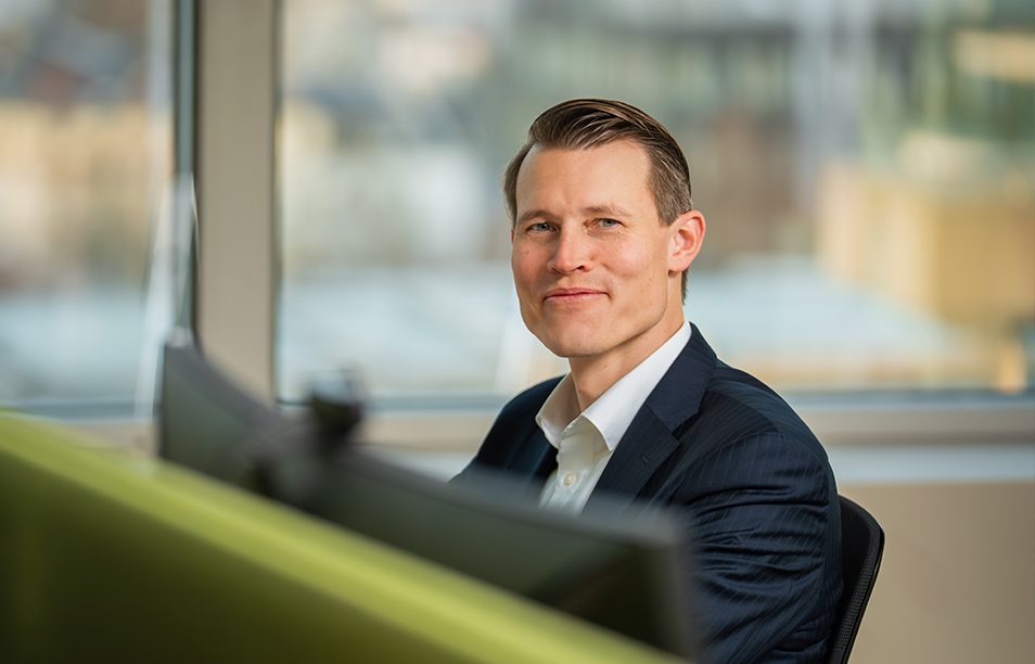 Das Bild zeigt eine Person in einem Büro oder Arbeitsplatz. Die Person sitzt vor mehreren Bildschirmen und trägt einen dunklen Anzug mit einem weißen Hemd. Im Hintergrund sind Fenster mit einem unscharfen Ausblick zu sehen.