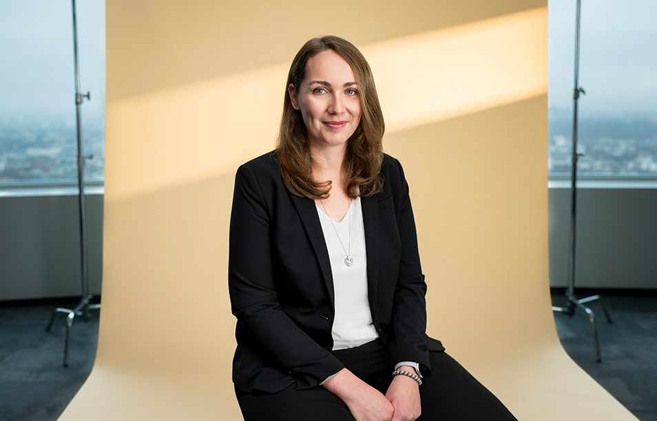 Das Bild zeigt eine Person, die vor einem beigen Hintergrund sitzt. Die Person ist in formeller Kleidung gekleidet, mit einem schwarzen Blazer, einem weißen Hemd und einer schwarzen Hose.