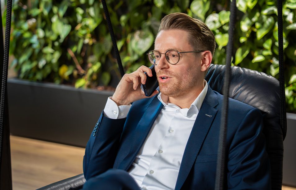 Das Bild zeigt eine Person in einem blauen Anzug, die auf einem Sessel sitzt und ein Handy ans Ohr hält. Im Hintergrund ist eine Wand mit Pflanzen zu sehen.