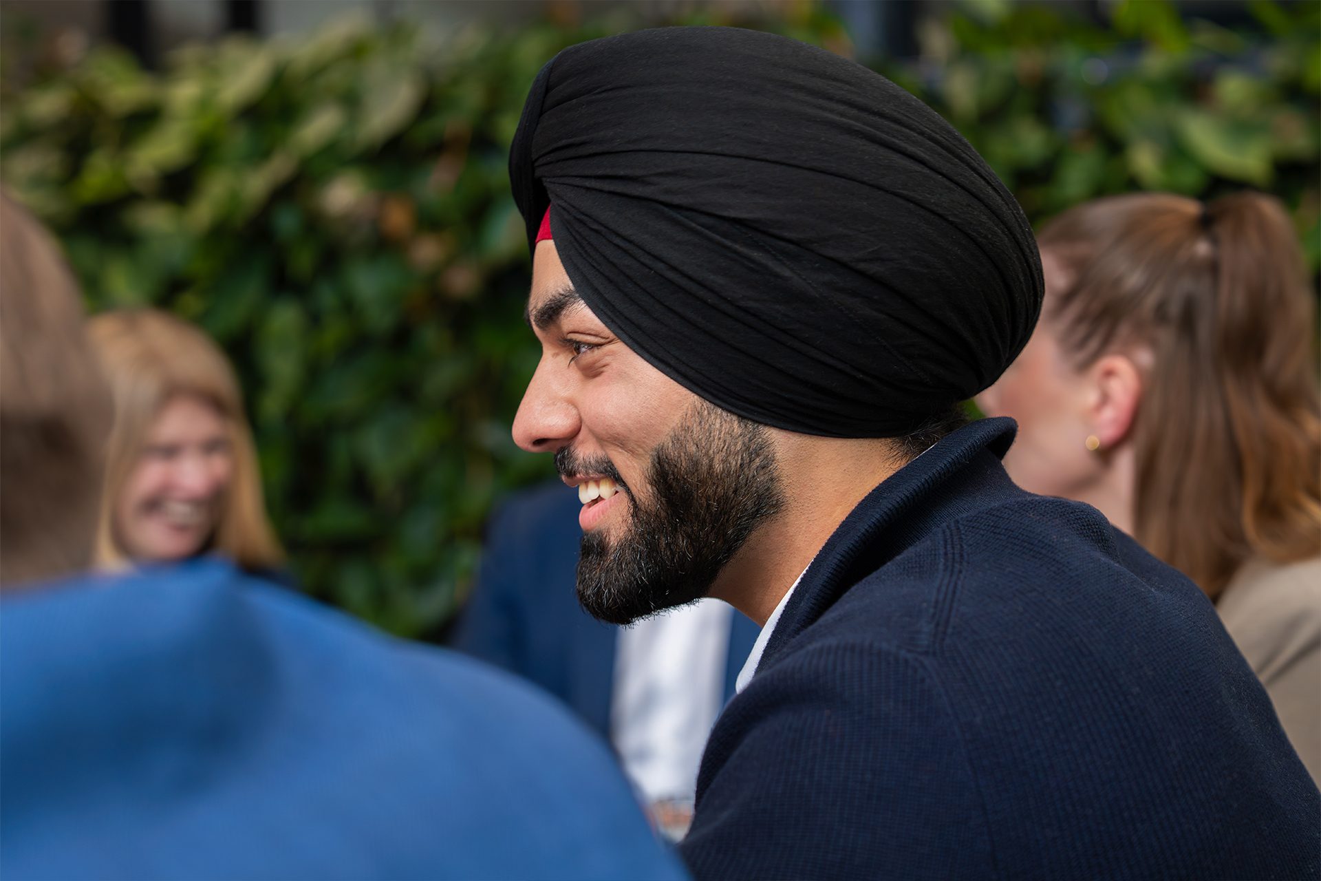 Das Bild zeigt eine Gruppe von Menschen, die sich unterhalten. Einer der Menschen trägt einen Turban. Der Hintergrund besteht aus grünem Laubwerk.