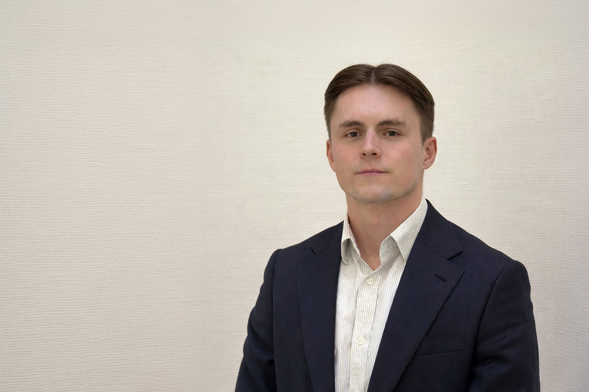 Das Bild zeigt eine Person in einem dunklen Anzug mit weißem Hemd. Der Hintergrund ist beige und die Person steht frontal zur Kamera. Das Gesicht ist unscharf gemacht, wodurch die Identität der Person nicht erkennbar ist. Es gibt keine weiteren Gegenstände oder Details im Bild.