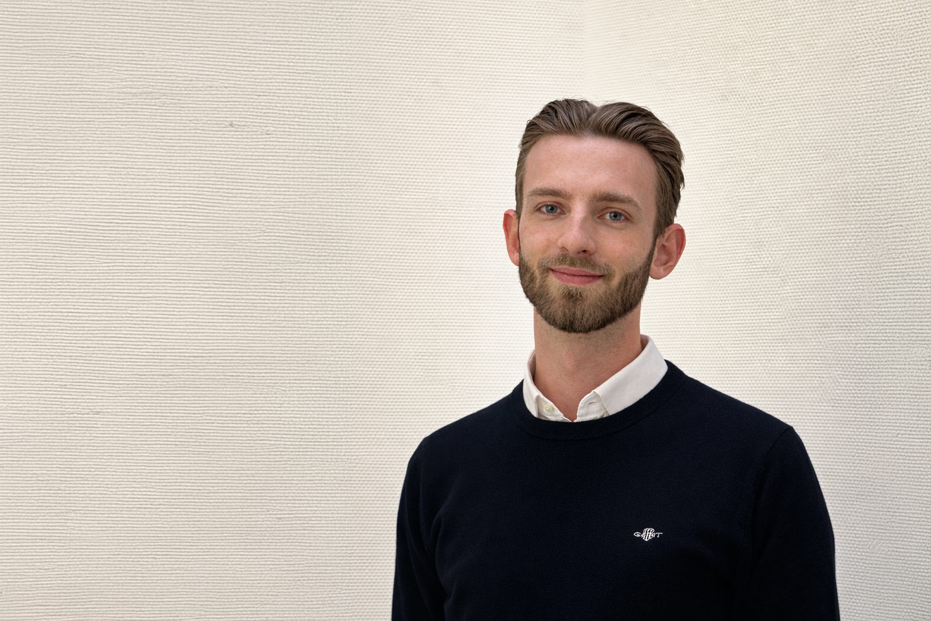 Das Bild zeigt eine Person, deren Gesicht verpixelt ist. Die Person trägt einen dunklen Pullover über einem weißen Hemd und steht vor einem hellen Hintergrund.